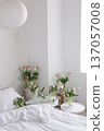 Multiple pink tulip bouquets in glass vases arranged around white bed in soft daylight. Fresh spring flowers creating airy minimalist bedroom atmosphere. 137057008