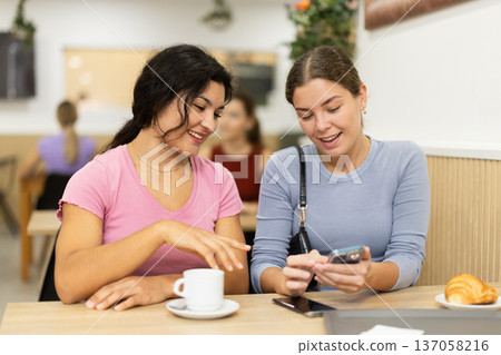 Latin girl is chatting with female friend at table in cafe, showing photos on phone screen Latin girl is chatting with female friend at table in cafe, showing photos on phone screen 137058216