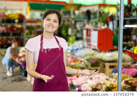 Portrait of smiling young woman gardener standing at greenhouse with flowers on background 137058374
