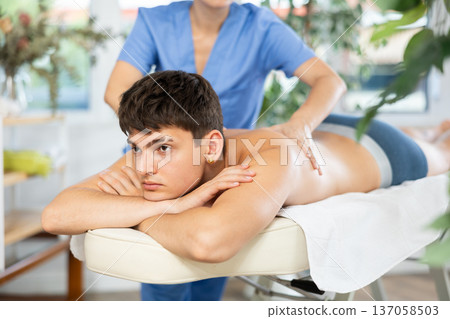 Female masseur doing massage at back man who lying on massage table in beauty salon 137058503