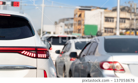 Rear side of luxury white car with turn on brake light. Many car on the road stop on traffic red light control. Blurred background of residential buildings in the city. Environment at day time. Rear side of luxury white car with turn on brake light. Many car on the road stop on traffic red light control. Blurred background of residential buildings in the city. Environment at day time. 137058676