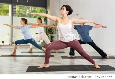 Active young Latin woman practicing Virabhadrasana 2 Warrior pose, indoor class with women, studio background Active young Latin woman practicing Virabhadrasana 2 Warrior pose, indoor class with women, studio background 137058866