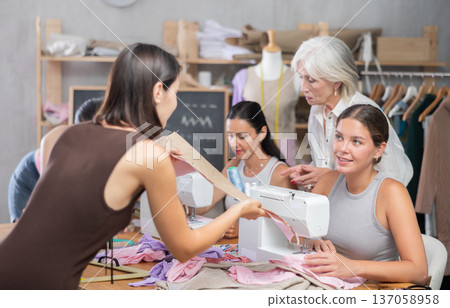 Girl stitching at machine and talking to groupmate in sewing workshop Girl stitching at machine and talking to groupmate in sewing workshop 137058958