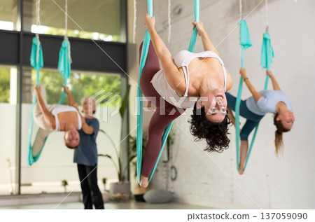 Fit young Latin woman doing fly yoga hanging upside down during female group training indoors in gym 137059090