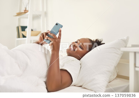 Smiling young woman waking up in bed in modern bedroom and using mobile phone. Smiling young woman waking up in bed in modern bedroom and using mobile phone. 137059836
