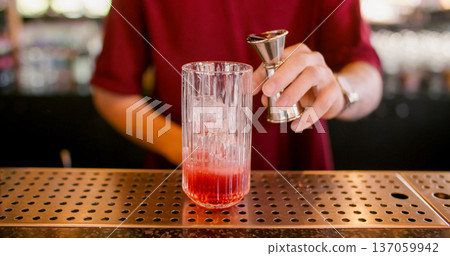 Cocktail making. Bar service. Bartender using jigger pouring red liquid to glass. Refreshing drink with ice cubes on metal counter. 137059942