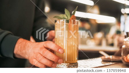 Cold drink. Served cocktail. Bartender hand offering tall glass of juice ice cubes with aromatic sprig green mint dried orange slice. 137059948