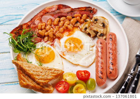 A classic breakfast plate featuring sunny-side-up eggs, crispy bacon, sausages, and fresh tomatoes A classic breakfast plate featuring sunny-side-up eggs, crispy bacon, sausages, and fresh tomatoes 137061801