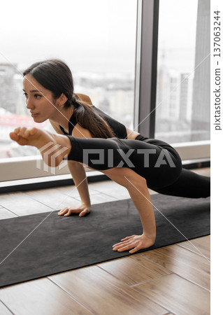 A woman in black activewear demonstrates a challenging yoga pose on a mat in front of a large window with a city view 137063244