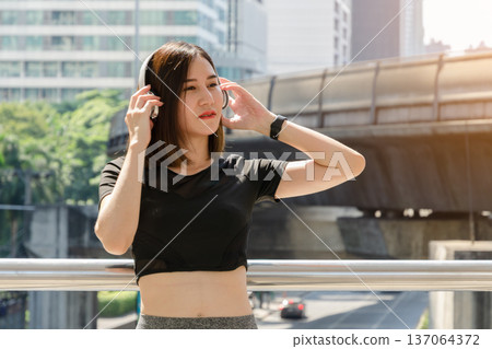 Young Asian woman with brown hair and dressed in athletic wear enjoying music through white headphones while standing on urban bridge or walkway Young Asian woman with brown hair and dressed in athletic wear enjoying music through white headphones while standing on urban bridge or walkway 137064372