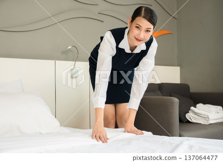 Smiling hotel Asian maid in dark blue uniform making bed with white linens in modern guest room Smiling hotel Asian maid in dark blue uniform making bed with white linens in modern guest room 137064477