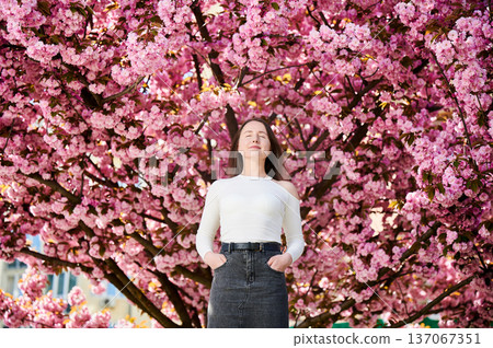 Woman allergic enjoying after treatment from seasonal allergy at spring. Portrait of happy beautiful woman with eyes closed in front of blooming sakura tree at springtime. Spring allergy concept. 137067351