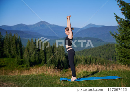 Woman practicing yoga outdoors in the mountains in a serene, natural setting. Female performing yoga pose on mat, with backdrop of beautiful mountain landscape at sunrise or sunset. Woman practicing yoga outdoors in the mountains in a serene, natural setting. Female performing yoga pose on mat, with backdrop of beautiful mountain landscape at sunrise or sunset. 137067453