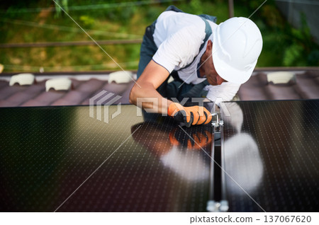 Worker building photovoltaic solar panel system on rooftop of house. Close up of man engineer in helmets and gloves installing solar module with help of hex key outdoors. Alternative, renewable energy Worker building photovoltaic solar panel system on rooftop of house. Close up of man engineer in helmets and gloves installing solar module with help of hex key outdoors. Alternative, renewable energy 137067620