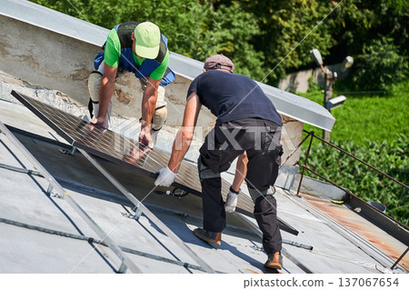 Workers building solar panel system on metal rooftop of house. Two men installers carrying photovoltaic solar module outdoors. Alternative, green and renewable energy generation concept. 137067654