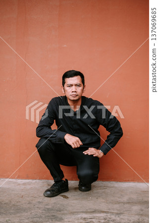 Serious young man in black squatting against a terracotta wall Serious young man in black squatting against a terracotta wall 137069685