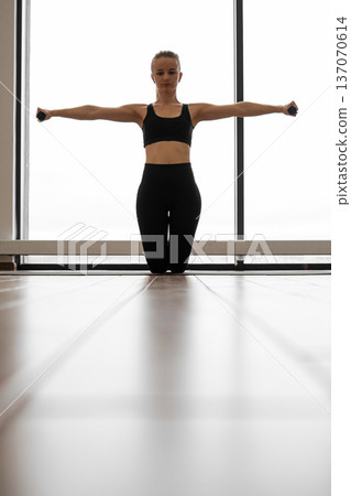 A woman in athletic wear kneels on a mat, performing lateral raises with dumbbells, her arms extended horizontally A woman in athletic wear kneels on a mat, performing lateral raises with dumbbells, her arms extended horizontally 137070614