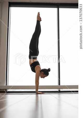 A woman in black athletic wear balances on her hands in a yoga studio with a large window behind her A woman in black athletic wear balances on her hands in a yoga studio with a large window behind her 137070633