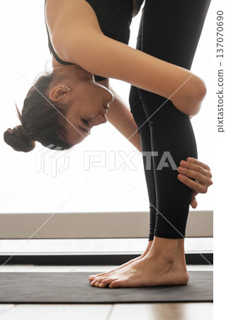 A woman in black activewear performs a standing forward fold yoga pose on a mat, emphasizing flexibility and balance A woman in black activewear performs a standing forward fold yoga pose on a mat, emphasizing flexibility and balance 137070690