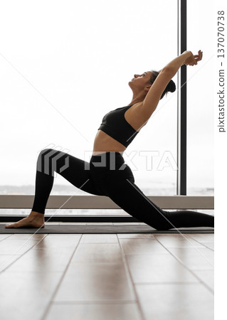 A woman in black activewear performs a crescent lunge yoga pose with her arms extended overhead, stretching her body A woman in black activewear performs a crescent lunge yoga pose with her arms extended overhead, stretching her body 137070738