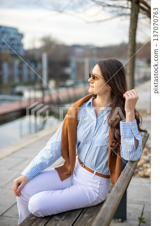 Elegant young woman in old money style sitting on bench near pier in Prague wearing sunglasses in early spring. Quiet luxury lifestyle and refined European elegance concept. 137070763