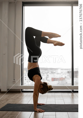 A woman performs a challenging yoga handstand pose on a mat in a studio with a large window overlooking a city skyline A woman performs a challenging yoga handstand pose on a mat in a studio with a large window overlooking a city skyline 137070766