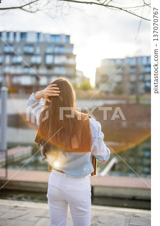 Elegant young woman in old money style standing by riverside in Prague during golden hour, back view portrait. Quiet luxury lifestyle and refined European femininity concept. Elegant young woman in old money style standing by riverside in Prague during golden hour, back view portrait. Quiet luxury lifestyle and refined European femininity concept. 137070767