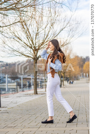 Elegant young woman in old money style walking along riverside promenade near pier in Prague during early spring. Understated luxury and timeless European lifestyle concept. Elegant young woman in old money style walking along riverside promenade near pier in Prague during early spring. Understated luxury and timeless European lifestyle concept. 137070770