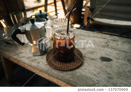 Refreshing iced black coffee in a glass with moka pot on a rustic wooden table. Refreshing iced black coffee in a glass with moka pot on a rustic wooden table. 137070909
