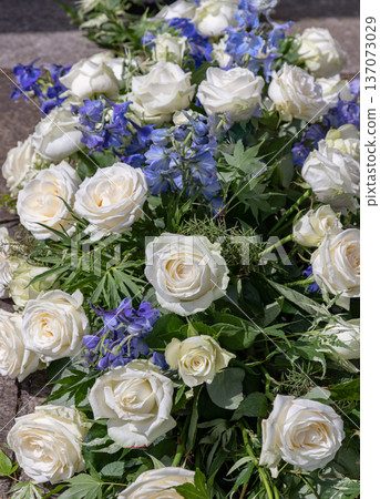 Full-bodied white roses and spires of cornflower delphinium are elegantly interwoven against a textured backdrop of fern and maple foliage, creating a luminous display of creamy petals and vibrant 137073029