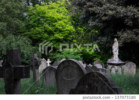 At Highgate Cemetery, a statue of Jesus overlooks a collection of weathered stone monuments and tall grass. The scene features a prominent Celtic cross on the left and a small white cross partially 137073180