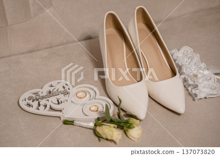 Elegant White Wedding Shoes with Lace Garter, Heart-Shaped Ring Holder, and Delicate White Roses on Beige Fabric Background Elegant White Wedding Shoes with Lace Garter, Heart-Shaped Ring Holder, and Delicate White Roses on Beige Fabric Background 137073820