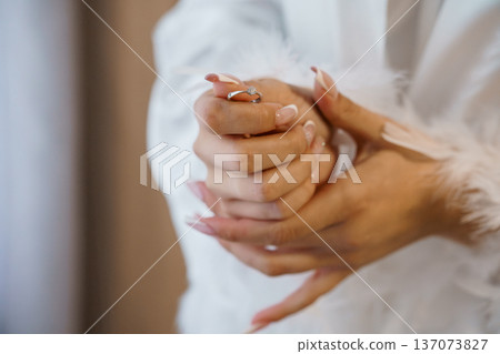 Close-up of a woman's hands holding a diamond engagement ring with feathered white robe in soft natural light Close-up of a woman's hands holding a diamond engagement ring with feathered white robe in soft natural light 137073827