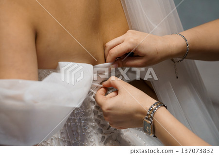Close-up of bride adjusting white wedding dress corset laces with hands, delicate beaded details and soft tulle veil in elegant bridal preparation 137073832