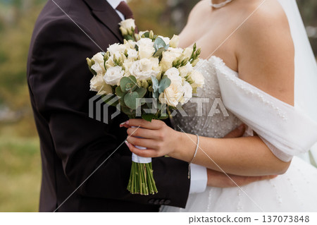 Elegant wedding couple embracing outdoors with white rose bouquet and lace dress in natural daylight Elegant wedding couple embracing outdoors with white rose bouquet and lace dress in natural daylight 137073848