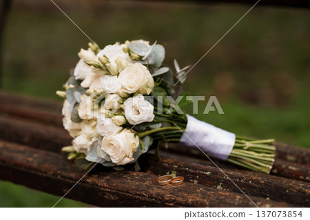 Elegant White Wedding Bouquet with Eucalyptus and Gold Rings on Rustic Wooden Bench in Natural Outdoor Setting 137073854