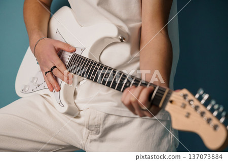 Close-up of a person playing a white electric guitar with black fretboard against a teal background 137073884