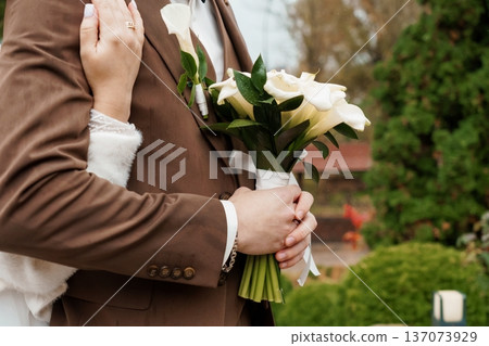 Elegant Wedding Couple Embracing Outdoors with White Calla Lilies and Brown Suit in Natural Garden Setting 137073929
