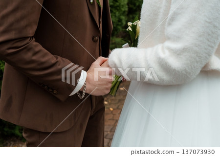 Close-up of bride and groom holding hands in wedding attire with white bouquet and brown suit against green garden backdrop Close-up of bride and groom holding hands in wedding attire with white bouquet and brown suit against green garden backdrop 137073930