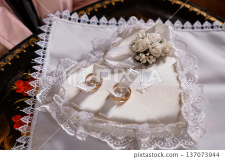 Elegant White Wedding Ring Pillow with Lace Trim and Pearl-Adorned Bands on Ornate Black Background Elegant White Wedding Ring Pillow with Lace Trim and Pearl-Adorned Bands on Ornate Black Background 137073944