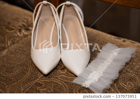 White Wedding Shoes with Tulle Garter on Ornate Fabric Background, Elegant Bridal Accessories in Soft Lighting White Wedding Shoes with Tulle Garter on Ornate Fabric Background, Elegant Bridal Accessories in Soft Lighting 137073951