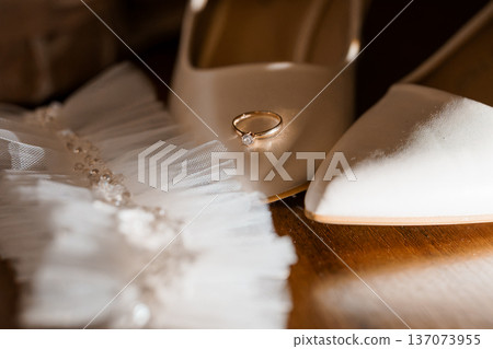 Elegant Wedding Ring Resting on White Bridal Shoes with Tulle Garter on Wooden Floor Elegant Wedding Ring Resting on White Bridal Shoes with Tulle Garter on Wooden Floor 137073955