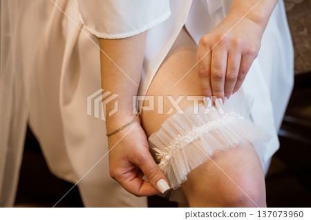 Bride wearing white lace garter during wedding preparations, close-up of hands adjusting tulle accessory on leg Bride wearing white lace garter during wedding preparations, close-up of hands adjusting tulle accessory on leg 137073960