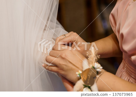 Close-up of a bridesmaid adjusting a bride's veil during wedding ceremony with soft pink and white tones and delicate floral wrist corsage Close-up of a bridesmaid adjusting a bride's veil during wedding ceremony with soft pink and white tones and delicate floral wrist corsage 137073965
