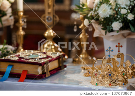 Golden Wedding Crowns and Ornate Religious Book on Altar with White Flowers and Candlelight in Church Interior 137073967
