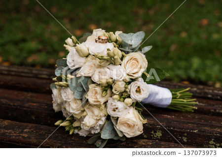 Elegant White Wedding Bouquet with Gold Rings on Wooden Bench in Natural Outdoor Setting Elegant White Wedding Bouquet with Gold Rings on Wooden Bench in Natural Outdoor Setting 137073973