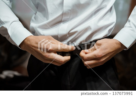 Close-up of a man adjusting a black belt over a white dress shirt, showcasing formal attire and meticulous grooming Close-up of a man adjusting a black belt over a white dress shirt, showcasing formal attire and meticulous grooming 137073991