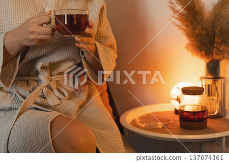 Woman in bathrobe holding a cup of tea, unwinding in a warm, comfortable home environment cozy relaxing for an evening or morning 137074361