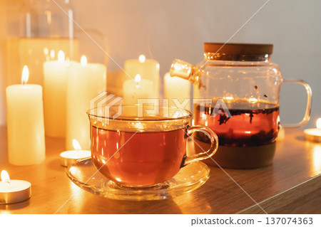 Steaming tea in a clear glass cup and teapot surrounded by flickering candles, creating a warm, cozy, and relaxing ambiance, creating hygge atmosphere 137074363