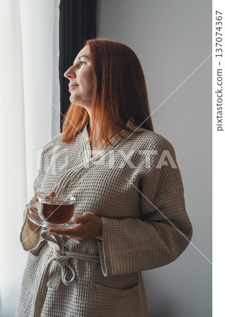 Woman in bathrobe drinking hot tea by window, finding peace and relaxation at home during a tranquil morning routine Woman in bathrobe drinking hot tea by window, finding peace and relaxation at home during a tranquil morning routine 137074367